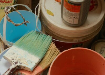Several paint buckets, a large orange mixing jug, a blue container, and two paintbrushes with green and white bristles sit stacked in a cluttered corner—tools ready for any Upstate New York painting contractor tackling interior painting projects.