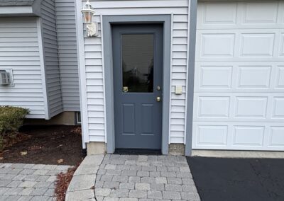 A gray exterior door with a window is set in a light gray vinyl-sided house, freshly finished by a trusted painting company Upstate New York; a curved stone walkway leads to the door, with a lamp fixture mounted above.
