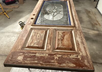 A large wooden door with a decorative glass window and two lower panels lies flat on a tarp in a garage—prepped by an Upstate New York painting contractor. Tools and ladders are visible, ready for expert exterior painting Upstate New York homeowners trust.